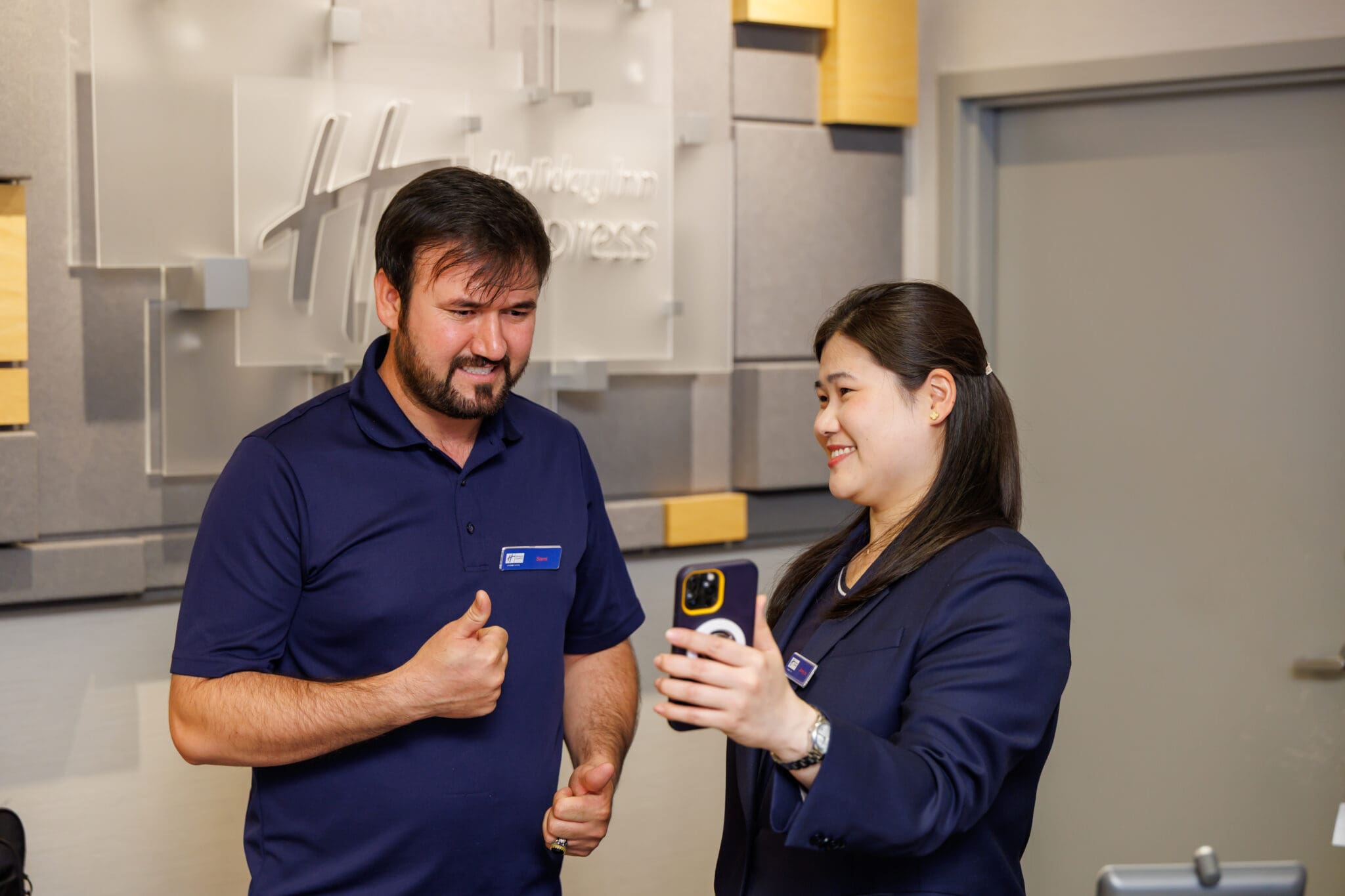 At the Holiday Inn Express front desk, a male staff member in a navy shirt uses sign language while a female colleague in a navy blazer holds up a smartphone showing a live interpreter. This moment is part of a Wavefront Centre and Holiday Inn Express Metrotown pilot of Virtual On-Demand Interpreting, enabling real-time communication between Deaf and hearing individuals without advance booking.